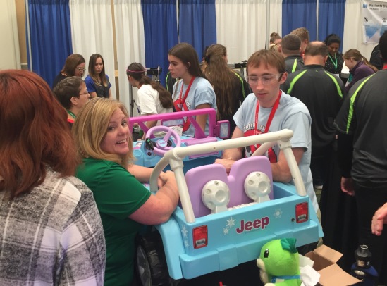 A young man and woman working on a toy Jeep in a busy exhibit hall.