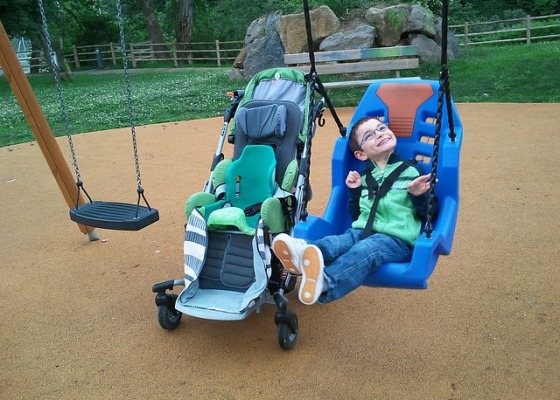 A little boy in an accessible playground swing next to his specialized stroller smiles at the sky.