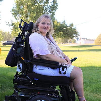 A woman smiling seated in her power wheelchair outside on the grass.