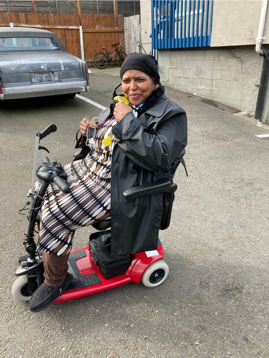 A middle-aged Black woman smiles outside seated in her power scooter